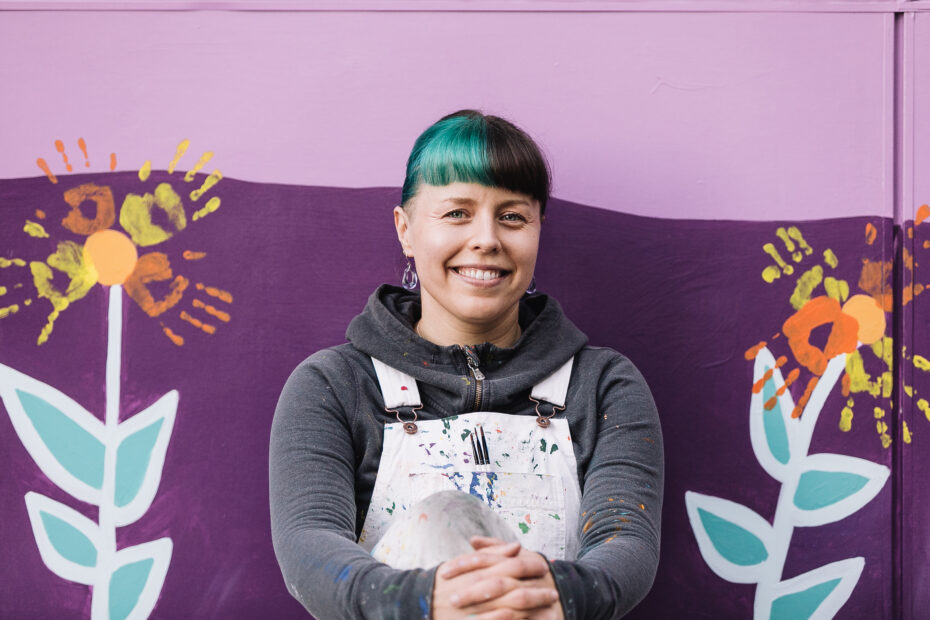 Artist Aistė Rye sitting in front of the mural she designed for the Northwest School for Deaf and Hard-of-Hearing Children. Aistė is sitting in front of of flowers that are made with the painted handprints of children who helped her paint the mural. .